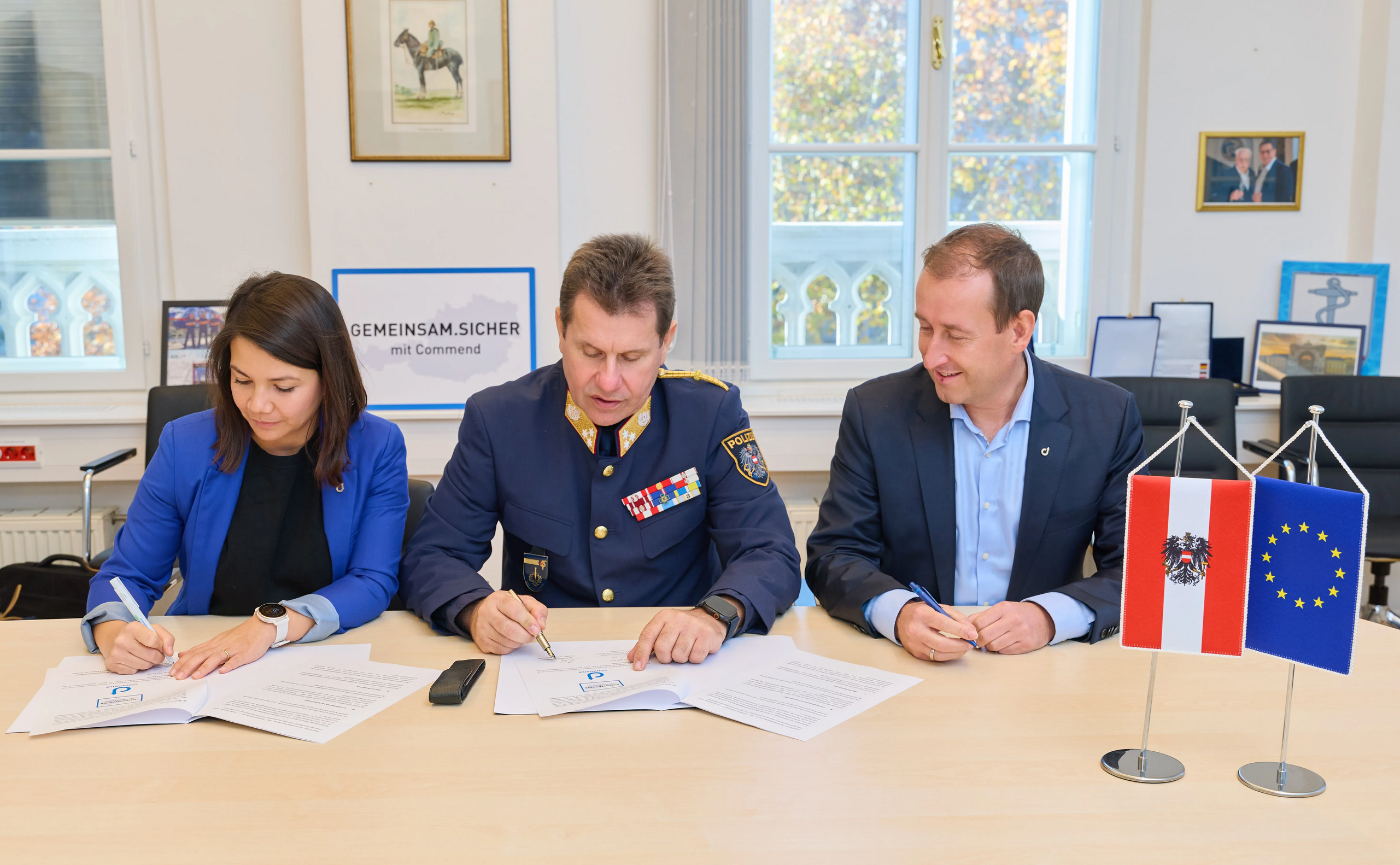 Security Partnership Agreement People sign documents at a table with flags; police officer present. Commend security or intercom solution.