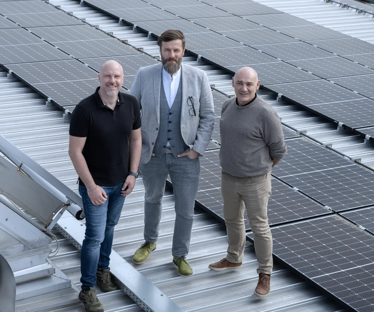 Partenaires commerciaux sur toit solaire Trois hommes, dont un barbu, sourient sur un toit ensoleillé avec de nombreux panneaux solaires.