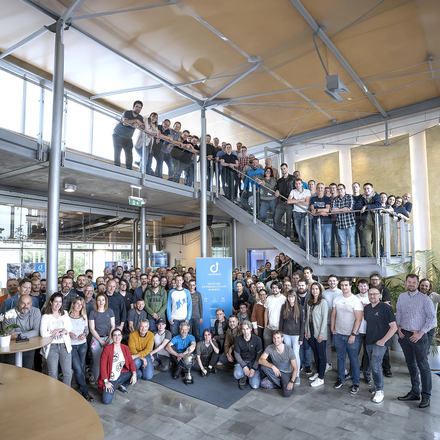 L'équipe Commend en photo de groupe Grand groupe d'employés de Commend dans un bureau moderne, posant pour une photo d'équipe avec un trophée.