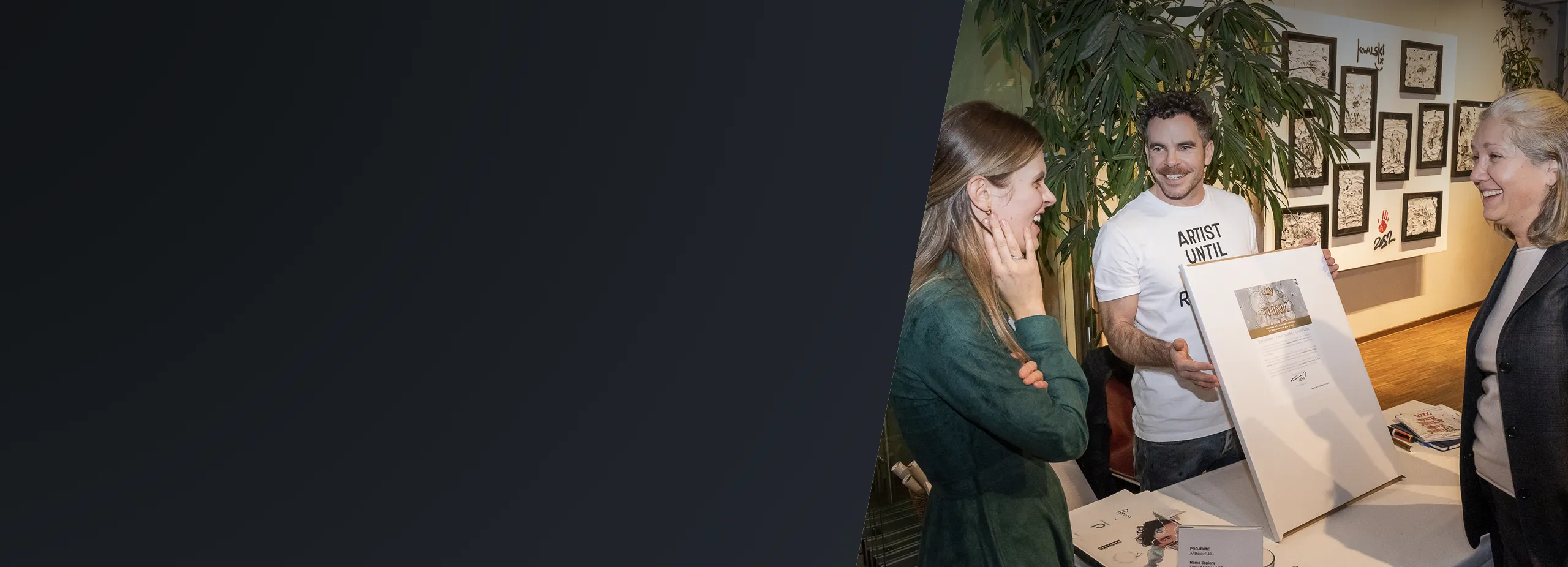 Joyful Art Presentation at Gallery Event Man presenting framed art to two smiling women at a gallery, with artworks on display.